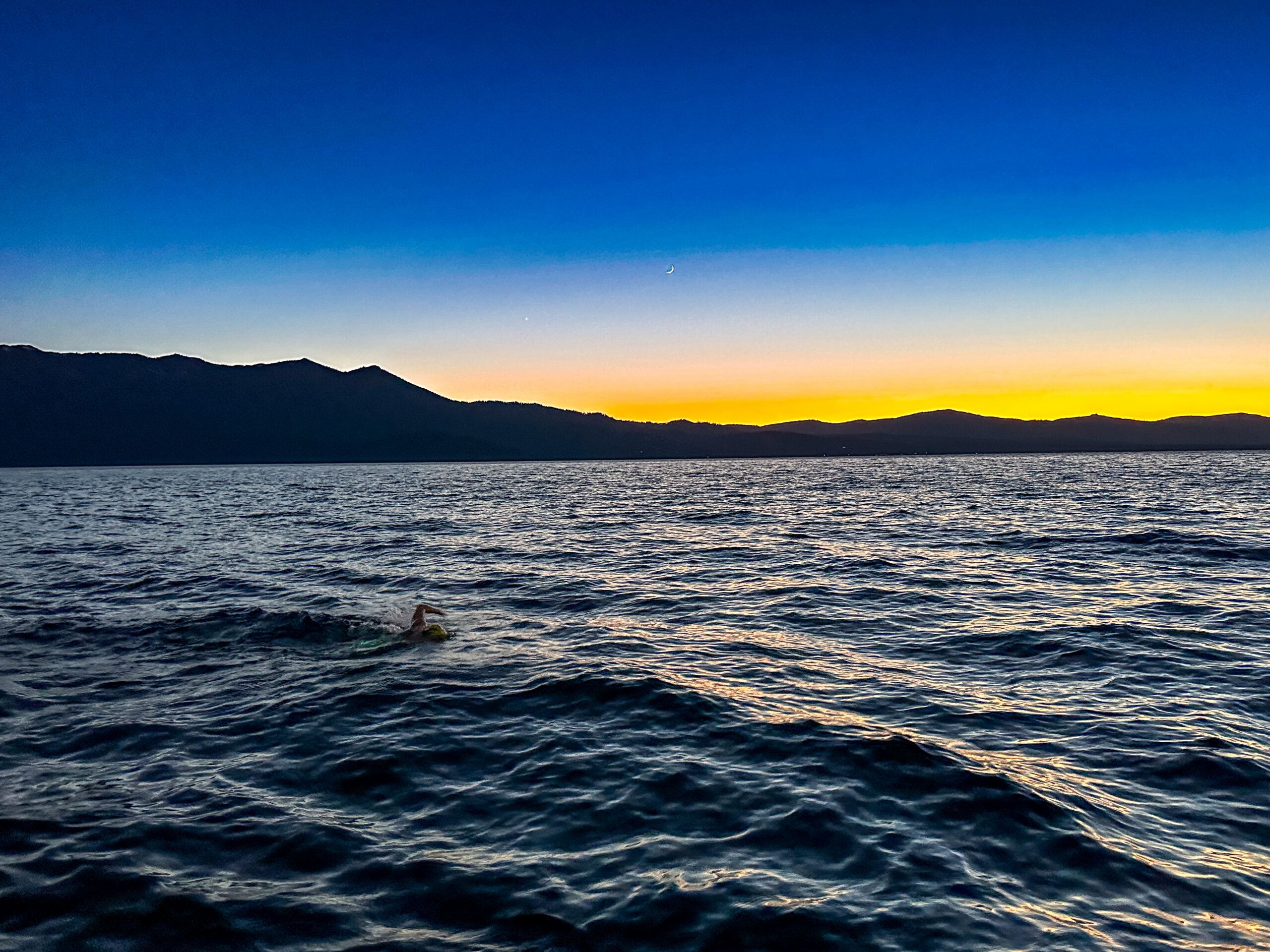A swimmer crosses Lake Tahoe beneath a crescent moon and a golden mountain horizon.